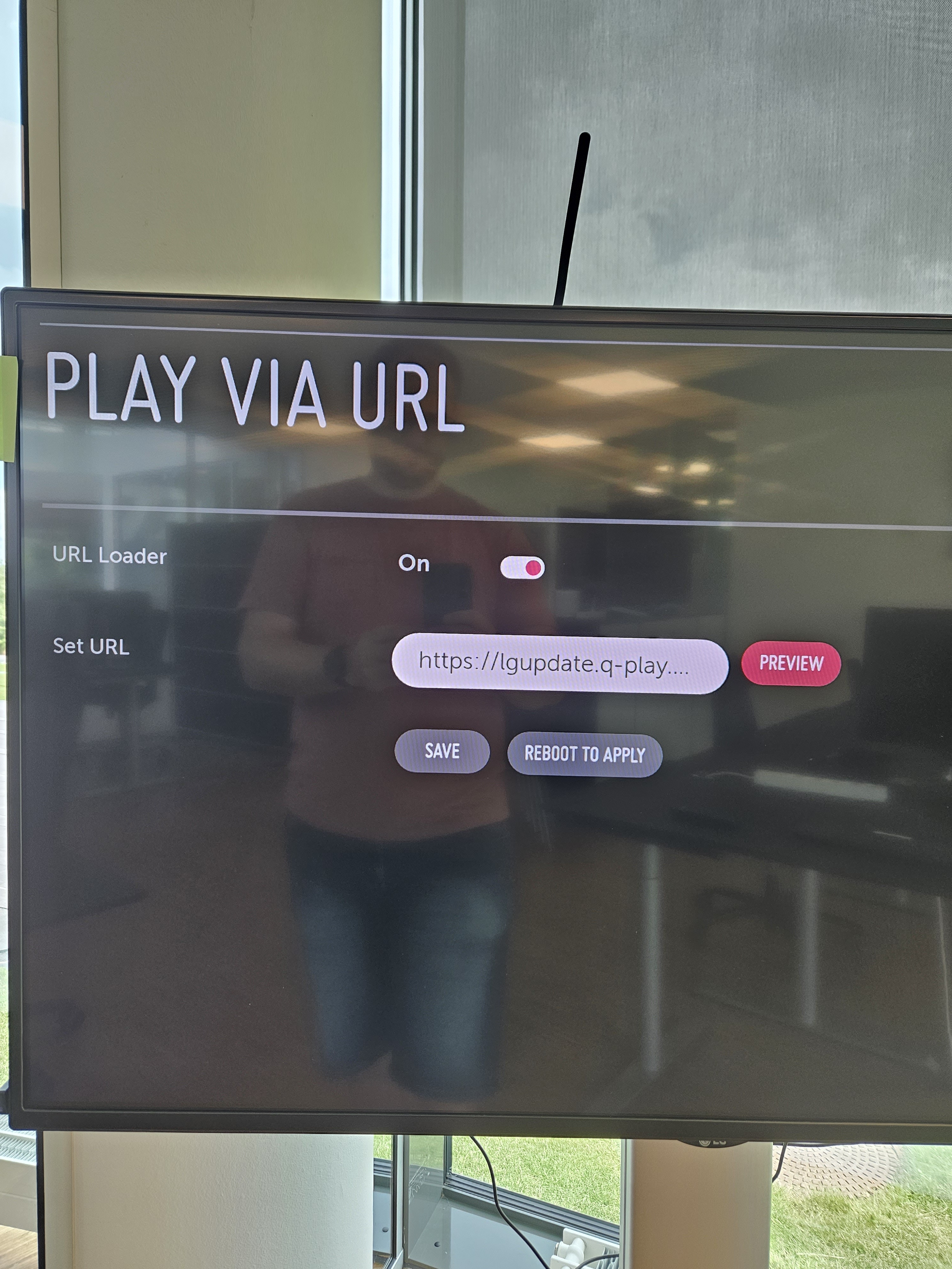 Digital display interface titled 'PLAY VIA URL' with toggles for URL Loader set to On, input field for URL, and buttons for Save, Preview, and Reboot to Apply.